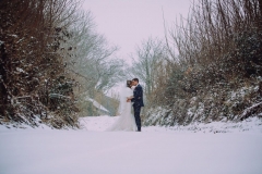 A  full length shot of the bride and groom standing embracing in the snow.. They are standing side by side and facing towards each other. The bride is on the left. The bare winter trees are towering above them. The bride is wearing a white cap sleeved dress . She has dark hair that she is wearing loose with a veil. . The groom is wearing a dark grey suit