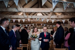 A bride walking down the "aisle" in the wedding barn  with her dad. The bride is on the left and they are linking arms. The bride is carrying a bouquet in her right hand. She is wearing a white cap sleeved dress . She has dark hair that she is wearing loose with a veil. You can see bunting and beams above their heads. The guests are standing and facing forward but turning their heads towards the bride and her father