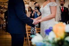 A close up shot of the bride and groom for the knees to their necks  in the wedding barn while they exchange vows. There are some flowers in the foreground