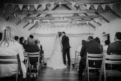 A black and white shot of the couple embracing at the front of the wedding barn after they have exchanged their vows. You can see beams and bunting above their heads. Their guests are seated on wooden chairs on either side of the "aisle"