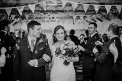 A black and white shot of the couple leaving the wedding barn after the ceremony.  They are walking down the "aisle" in the wedding barn  arm in arm. The bride is on the right. The bride is carrying a bouquet in her left hand. She is wearing a white cap sleeved dress . She has dark hair that she is wearing loose with a veil. You can see bunting and beams above their heads. The guests are standing and facing forward but turning their heads towards the bride and her groom.