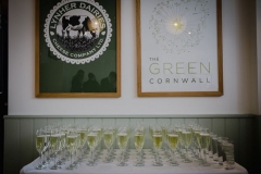 An image of charged glasses of champagne set up on a clothed white table. Above the table there are 2 framed pictures on the wall. One is a vintage advert for Yarg cheese and the other is the logo of The Green.