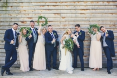 This image shows the bridal party including the bride and groom, 3 bridesmaids and 3 groomsmen. The group is standing on a lin (boy, girl, boy, girl etc. ) with the bride and groom 5th and 6th from the left. The  bridesmaids are holding greenery hoops which they have as an alternative to bouquets. This is a light hearted and informal shot with the bridesmaids looking through the hoops and waving them in the air. The groom and groomsmen wear navy suits, gray waistcoats, white shirts and blue ties. The bride wears  a full-length, sleeveless, white dress with lace details  and a beaded bodice. The bridesmaids wear pale pink dresses similar in style to the bride's.
