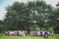 This is an image of the Oak Arbour just before the couple arrive. The arbour is a wooden structure with four corner posts supporting a pitched roof. The arbour has a large Oak Tree on each side. The trees are in full leaf. This photo is taken from a bit of a distance and the whole of the Oak Trees are in the frame. There is a clothed table (white) also under the arbour.  It is a summers day and the trees are in full leaf. You can see their guests sitting in front of the arbour in two columns of rows of seats.. The guests are looking at the couple towards the arbour and have their backs to the camera. You can see  a clothed table under the arbour.