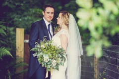 This is an image of the bride and groom standing in the woods. They are standing side by side with the groom on the left. The bride is angled slightly inwards towards the groom. The bride has a bouquet of greenery in her left hand.  The groom is wearing a navy suit, gray waistcoat, white shirt and blue tie. The bride wears  a full-length, sleeveless, white dress with lace details  and a beaded bodice. There is greenery in the background and a leafy branch in the foreground. There is some sheep fencing behind the couple.