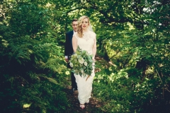 This is an image of the bride and groom walking in the woods. They are walking in single file on a narrow path with the groom behind. They are walking in a tunnel of greenery. The bride has a bouquet of greenery in her left hand.  The groom is wearing a navy suit, gray waistcoat, white shirt and blue tie. The bride wears  a full-length, sleeveless, white dress with lace details  and a beaded bodice.