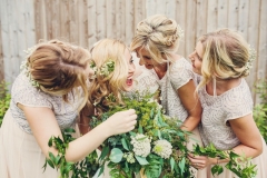 This image shows the bride and 3 bridesmaids The group is standing in a line in front of an oak fence. With the bride and groom 2nd from the left. The  bridesmaids are holding greenery hoops which they have as an alternative to bouquets. This is a light-hearted and informal shot with everyone laughing and bending in towards the bride. They are sharing a joke. All have blond hair.