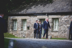 This is a photo the groom and his 3 groomsmen walking out of cottage number 1. The groom and groomsmen wear navy suits, gray waistcoats, white shirts and blue ties. Cottage number one sleeps two people. The property is single storey. The photo shows the full width of the cottage. In the background you can see trees which are in leaf. In the foreground you can see two patches of grass one on the left and one on the right and in the front there is gravel. The cottage is made of granite stone and is brown and grey in colour. The roof is constructed of grey slate and is pitched with the roof line running from left to right of the photograph along the length of the cottage. In the middle of the front of the cottage there is a wooden door that is painted pale green on either side of the story there are two windows on each side of this door. They are Georgian style sash windows and also painted green. On the right hand side of the cottage the gable end has hanging slate on it. There is a French window in this end of the cottage and it is also painted green. In the foreground of the picture there is a light bollard for outside lighting.