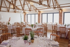 This is an image of the Red Brick barn set up for a wedding meal. The room has oak floors and white walls and ceiling. The tables are round and covered in white table cloths. There are bi-fold doors to the front of the room away from the camera. Light is streaming in the windows. There are wooden (cheltenham-style) chirs around the tables. Youc an see 5 tablesin the picture. The tables are laid up with white crockery and silverware. There are pots of green plants in the centreof each table as centre pieces. There are no people in the room yet.