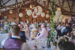 This is an image of the Red Brick barn during a wedding meal. The room has oak floors and a red brick wall and white ceiling. The tables are round and covered in white table cloths. There are wooden (cheltenham-style) chirs around the tables. You can see around 5 tables in the picture. The tables are laid up with white crockery and silverware. There are pots of green plants in the centreof each table as centre pieces. There are people seated at the tables. There are balloons rising up towards the A-frame beamsin the pitched roof.