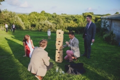 A group of guests are playing Giant Jenga on a green lawn, The shadows are long in this shot indicating that it is early evening. There are a group of 4 guests playing the game. In the background there are many green trees.