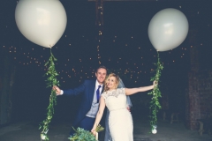 This is an image of the bride and groom standing in the doorway of the Really Rustic barn. They are standing side by side with the groom on the left. The bride is leaning slightly inwards towards the groom. The bride has a bouquet of greenery in her right hand. The couple each have their outside arms outstretched and each is holding  a very large white balloon tethered by a "string" of greenery. The groom is wearing a navy suit, gray waistcoat, white shirt and blue tie. The bride wears  a full-length, sleeveless, white dress with lace details  and a beaded bodice.