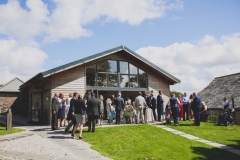 This is an image of our Green Room Bar. The building is clad in oak planks. It has a pitched roof and glazing at the front. It has bi-fold doors that are open. Guests are spilling out of the doors onto the lawn in front of the building. The sky behind is bright blue with some clouds. There is a granite chippin path leading up to the building to the left of the shot. Ther are aound 30 weddnig guests visible in the picture.