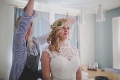 This is an image of the bride getting ready. She is seated in a chair in the dining room of the house. She is facing to the front but angled slighly to the right of the camera. There is someone behind her and they are positioing her veil. The assistant is wearing a striped shirt and a dark apron. The bride wears  a  sleeveless, white dress with lace details  and a beaded bodice. The curtains and walls in the background are pale blue and the curtains are closed. The other wall and ceiling are painted white.