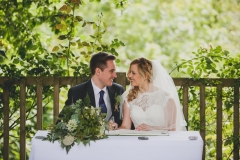 This picture shows an image of the bride and groom seated at the registrars' table just after they have exchanged their vows. They are seated side by side with the groom on the left. They are turned slightly towards each other and are looking in each others eyes. They are seated at a clothed white table and there is a bouquet of greenery on the left hand side of the table. . The groom is wearing a navy suit, gray waistcoat, white shirt and blue tie. The bride wears  a, sleeveless, white dress with lace details  and a beaded bodice. Their is a wooden fence and lots of greenery behind them.