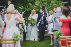 Newly weds leaving the Oak Arbour. The arbour is a wooden structure with four corner posts supporting a pitched roof. This photo shows a couple just walking away from the Oak Arbour after their ceremony. The couple are in the middle of the shot and the photo shows them in full length. They are holding hands and the groom is on the right, the bride on the left. The bride is wearing an white fitted dress with sequins and lace details.  She has  blond hair she is wearing loose. In her left hand she is holding a bouquet of green and cream flowers. The groom is wearing a dark suit with white shirt and a navy tie. The couple are smiling. You can see some guests around them. The guests are looking at the couple and clapping and have their backs to the camera. There are rows of chairs set out in front of the arbour but the guests are standing. There are "pew" flowers on the chairs on the "aisle" end of the rows.