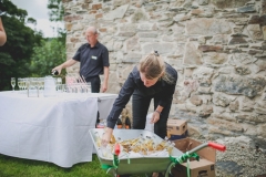 This picture shows 2 members of staff getting ready to serve drinks at an outside bar. They are setting up  in front a a stone wall that is made of gray and brown granite stones. There is a table clothed in a white table cover. One member of staff is settgin out glasses. Another is placing bottles of beer into a wheelbarrow that is full of ice. teh staff are both male and they are wearing black shirts and black trousers
