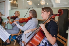 This shows a 3 musicians playing in The Green Room. They are paying strings. They are 3 women. tow are playing violins and one is playing a cello. They are seated in an a room that is painted pale  green. There are some sparkly lights and spot  lights in the background.