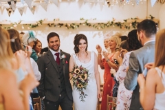 Amy and Kieran  leaving the weding barn hand in hand and smiling being cheered by their guests. Amy holds a bouquet of flowers