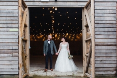The couple are standing in the doorway of the Really Rustic Barn. The doors are open and they are about twice the height of the couple. You can see festoon lighting in the backgorund inside the barn. The are facing the camera and holding hands.  The bride has She has dark hair worn up. She has a fitted white dress with a sweatheart neckline and lace sleeves. The groom is wearing a tweed 3 piece suit and brown bow-tie.