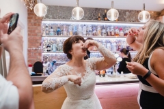 The bride is downing a shot  in the Red Brick Barn. You can see some guests and a bar in the backgorund inside the barn. She is facing the camera.  The bride has She has dark hair worn up. She has a fitted white dress with a sweatheart neckline and lace sleeves and a full skirt..