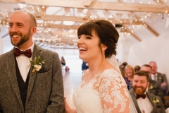 The bride and groom are facing the camera in the wedding barn during their ceremony. The bride has She has dark hair worn up. She has a fitted white dress with a sweatheart neckline and lace sleeves. The groom is wearing a tweed 3 piece suit and brown bow-tie.
