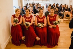 A group of 4 bridesmaids are seated in the front row of the left hand column of chairs inthe wedding barn, they are waring re dresses and holding flowers