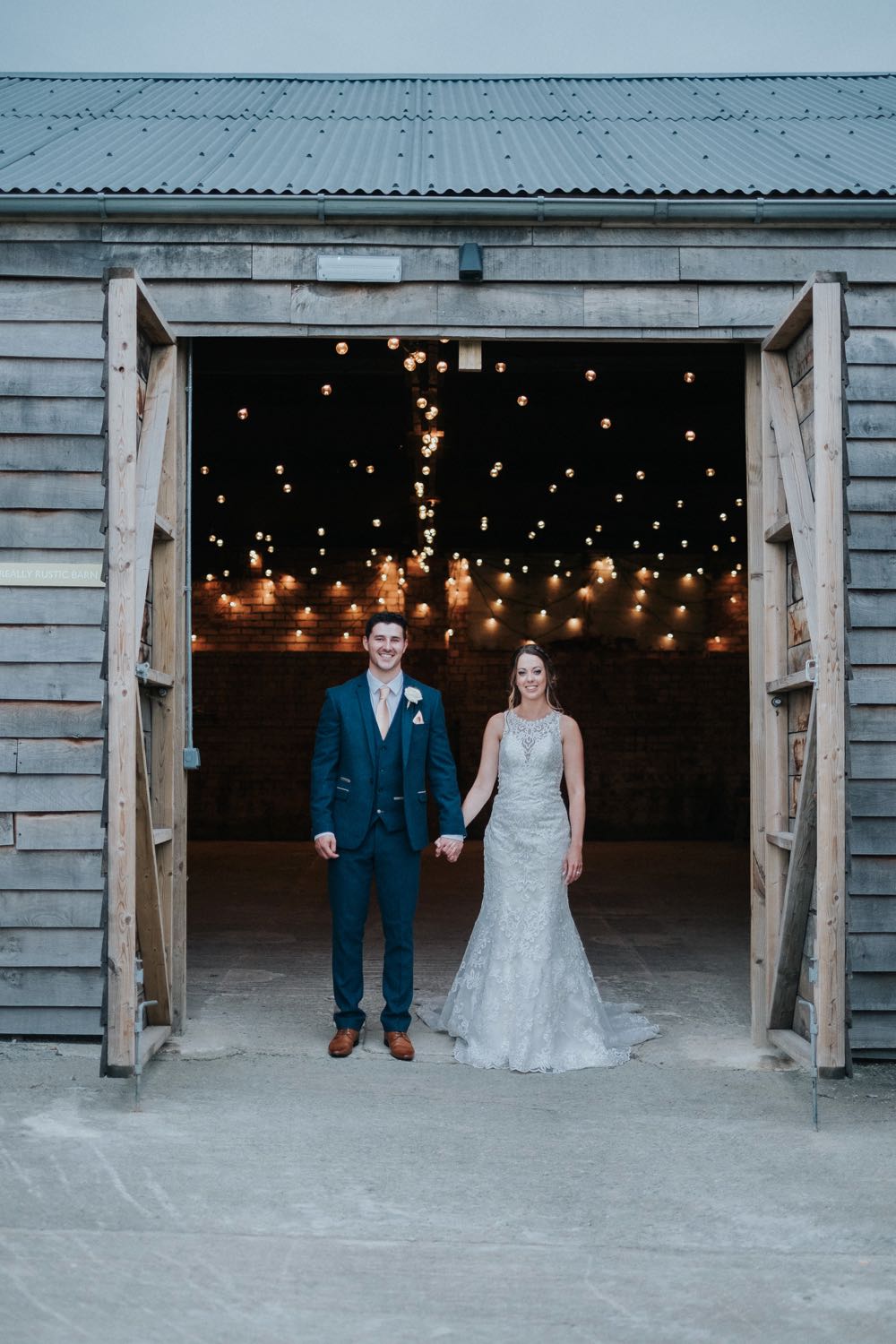 This picture is taken outside the really rustic barn. In the foreground facing the camera you can see the bride and groom. She is wearing a white, sleeveless  gown with a high neck and lace details. The groom is wearing  a dark suit, white shirt and light coloured tie. He has a white flower in his left lapel. The couple are standing side by side holding hands with the bride on the right and the groom on the left. tThe coule are facing the camera. You can see the festoon lighting in the really rustic barn ceiling.
