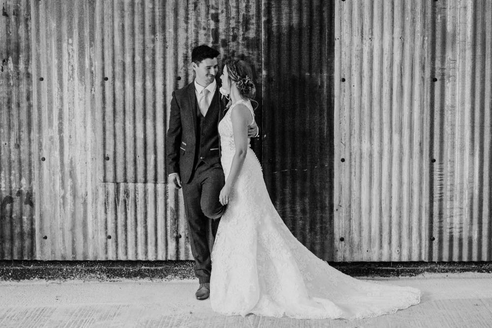 This black and white picture is taken in the Really Rustic Barns. It is a full length shot of the couple. She is wearing a white, sleeveless  gown with a high neck and lace details. The groom is wearing  a dark suit,  The couple are standing with the bride on the right and the groom on the left. They are facing each other.