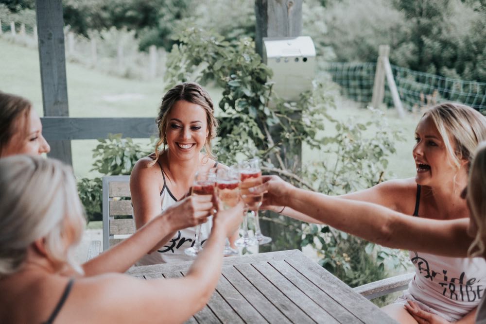 The bride surrounded by her 3 brodesmaids enjoys a drink on the patio. The four girls are seated and are clinking glasses with glasses raised. The bride is in the middle of the group.