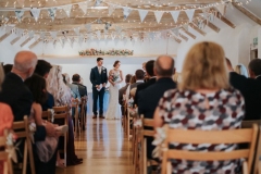 This picture is taken from the entrance end of the wedding barn. The guests  are seated in 2 columns of folding wooden chairs and they have their back to the camera.. At the other end of the barn, in the distance you can see the bride and groom. She is wearing a full length white gown and the groom is wearing  a dark suit. You can see the A frame beams inthe wedding barn roof with bunting strung across and the oak floor inthe aisle.