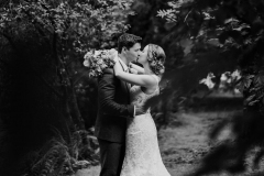 This black and white picture is taken outdoors. In the foreground facing each other and embracing you can see the bride and groom. She is wearing a white, sleeveless  gown with a high neck and lace details. She has a bouquet in her hand. The groom is wearing  a dark suit,  The couple are standing with the bride on the right and the groom on the left. They are facing each other and kissing. You can see lots of trees and bushes in the background.