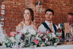 This picture shows the couple sitting on the top table during the wedding breakfast. The top table is set up in front of the red brick wall. the bride is on the right and the groom is on the left. The foreground is full of flowers.