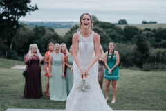 The bride is standing in the foreground facing the camera. She is wearing a white, sleeveless  gown with a high neck and lace details. Behind her onthe lawn there is a group of girls standing on a lawn who are waiting for the bride to throw her bouquet. In the background you can see trees and rolling hills. The lawns are parched giving the feeling that it is the height of summer.