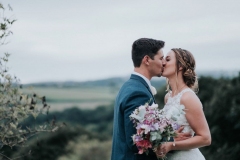 This picture is taken outdoors with rolling hills  in the background. In the foreground each other are the bride and groom. She is wearing a white, sleeveless  gown with a high neck and lace details. The groom is wearing  a dark suit, white shirt and light coloured tie.  The couple are standing  with the bride on the right and the groom on the left. They are facing each other and kissing. The bride has a large bouquet of white and pink flowers