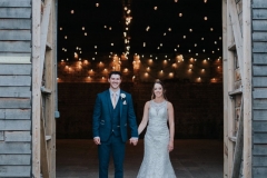 This picture is taken outside the really rustic barn. In the foreground facing the camera you can see the bride and groom. She is wearing a white, sleeveless  gown with a high neck and lace details. The groom is wearing  a dark suit, white shirt and light coloured tie. He has a white flower in his left lapel. The couple are standing side by side holding hands with the bride on the right and the groom on the left. tThe coule are facing the camera. You can see the festoon lighting in the really rustic barn ceiling.