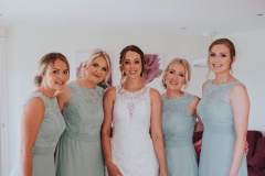 The bride is standing in her wedding dress flanked by 4 bridesmaids, 2 on each side of her. The bride is wearing a white sleevless gown with a high neck and lace details. The bridesmaids are waering gray dresses of a similar style. The photo is taken indoors.