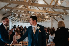 The groom is standing at the front (ceremony end) of the wedding barn. He as his back to the guests who are seated in 2 columns of folding wooden chairs behind him.  He is wearing a navy suit, white shirt and light coloured tie. He has a white flower in his left lapel. He is turning slightly to his right to shake hands with another man. The registrar is on his left and has her back to the camera. You can see all the A-frame roof beams of the weddin barn/