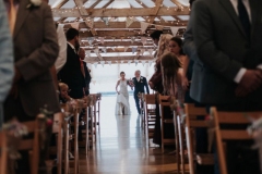 This picture is taken from the ceremony end of the wedding barn. The guests  are seated in 2 columns of folding wooden chairs. At the other end of the barn, in the distance you can see the bride coming into the barn escorted by her father. She is wearing a full length white gown and her faher is wearing a dark suit. You can see the a frame beams inthe wedding barn roof and the oak floor inthe aisle.