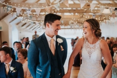 This picture is taken from the ceremony end of the wedding barn. The guests  are seated in 2 columns of folding wooden chairs and they are facing the camera in the background. In the foreground facing the camera you can see the bride and groom. She is wearing a white, sleeveless  gown with a high neck and lace details. The groom is wearing  a dark suit, white shirt and light coloured tie. He has a white flower in his left lapel. The couple are standing side by side with the bride on the right and the groom on the left. Their heads are turned slightly towards each other and they are laughing. You can see the A frame beams inthe wedding barn roof with bunting strung across and the oak floor inthe aisle.