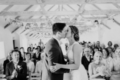 This black and white picture is taken from the ceremony end of the wedding barn. The guests  are seated in 2 columns of folding wooden chairs and they are facing the camera in the background. In the foreground facing the camera you can see the bride and groom. She is wearing a white, sleeveless  gown with a high neck and lace details. The groom is wearing  a dark suit, white shirt and light coloured tie. He has a white flower in his left lapel. The couple are standing side by side with the bride on the right and the groom on the left. They are facing each other and kissing. You can see the A frame beams inthe wedding barn roof with bunting strung across and the oak floor inthe aisle.