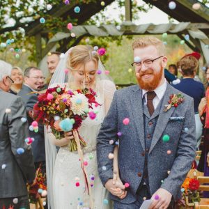 Newly weds leaving the Oak Arbour. The arbour is a wooden structure with four corner posts supporting a pitched roof. The front of the arbouris draped with cream voiles.This photo shows a couple just walking away from the Oak Arbour after their ceremony. Their guests are thowing pompom confetti. The couple are in the foreground and the shot shows them from the knees up. They are holding hands and the groom is on the right, the bride on the left. The bride is wearing a white fitted dress with lace details and lace cap sleeves. She has dark blond hair she is wearing. The bride is wearing a veil. In her left hand she is holding a bouquet of autumn coloured flowers: red, golds and browns flowers. The groom is wearing a grey, tweed, three-piece suit with a, white shirt and a brown tie. The groom has red hair, glasses and a beard. The couple are holding hands and the bride is looking down and smiling and the groom is looking off to his left and also smiling. You can see some guests around them. The guests are looking mostly at the couple but some have their backs to the couple. There are rows of chairs set out in front of the arbour but the guests are standing. You can see the registrar standing in the background under the arbour..