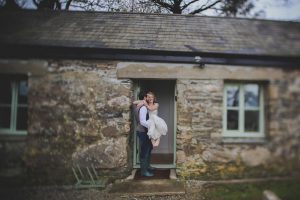 This is a photo of a groom carrying his bride over the threshold into cottage number 1. The groom is on the left, side on to the camera and the bride is on the right seated in the grrom's arms facing the camera. The groom has dar hair and is wearing dark trousers, a brown tweed waistcoat and a white shirt. He has a beard and is weraing green wellies. The brode is wearing a short-sleeved, full length white dress. She has light brown hair that she is wearing loose and her feet are bare. Cottage number one sleeps two people. The proerty is single storey. The photo shows part of the width of the cottage. In the background you can see trees which are bare. The cottage is made of granite stone and is brown and grey in colour. The roof is constructed of grey slate and is pitched with the roof line running from left to right of the photograph along the length of the cottage. The door is in the middle of the cottage and is painted pale green and is open. On either side of the door you can see a window. They are 4 pane casement style windows and are also painted pale green. On the left hand side of the door you can seee a 4 spoke welly rack painted pale green.