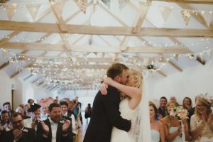 This photo shows the "you may kiss the bride" moment of a ceremony held in the wedding barn. In the foreground you can see the bride and groom having a kiss. The guests are seated behind the couple in two columns of rows of chairs. The wedding barn has an oak floor and the walls and ceiling are painted white. There is a vaulted ceiling with a sets of old, oak, a-frame beams supporting the roof. You can see 10 sets of the beams in the image. There are fairy lights garlanding the beams and there is bunting on the beams too. the guests are seated and are looking at the bride and groom and clapping. The groom is wearing a dark jacket and has light brown hair and a beard. The bride is wearing a lace, sleeveless dress and has blond hair worn loose. She is wearing a veil.