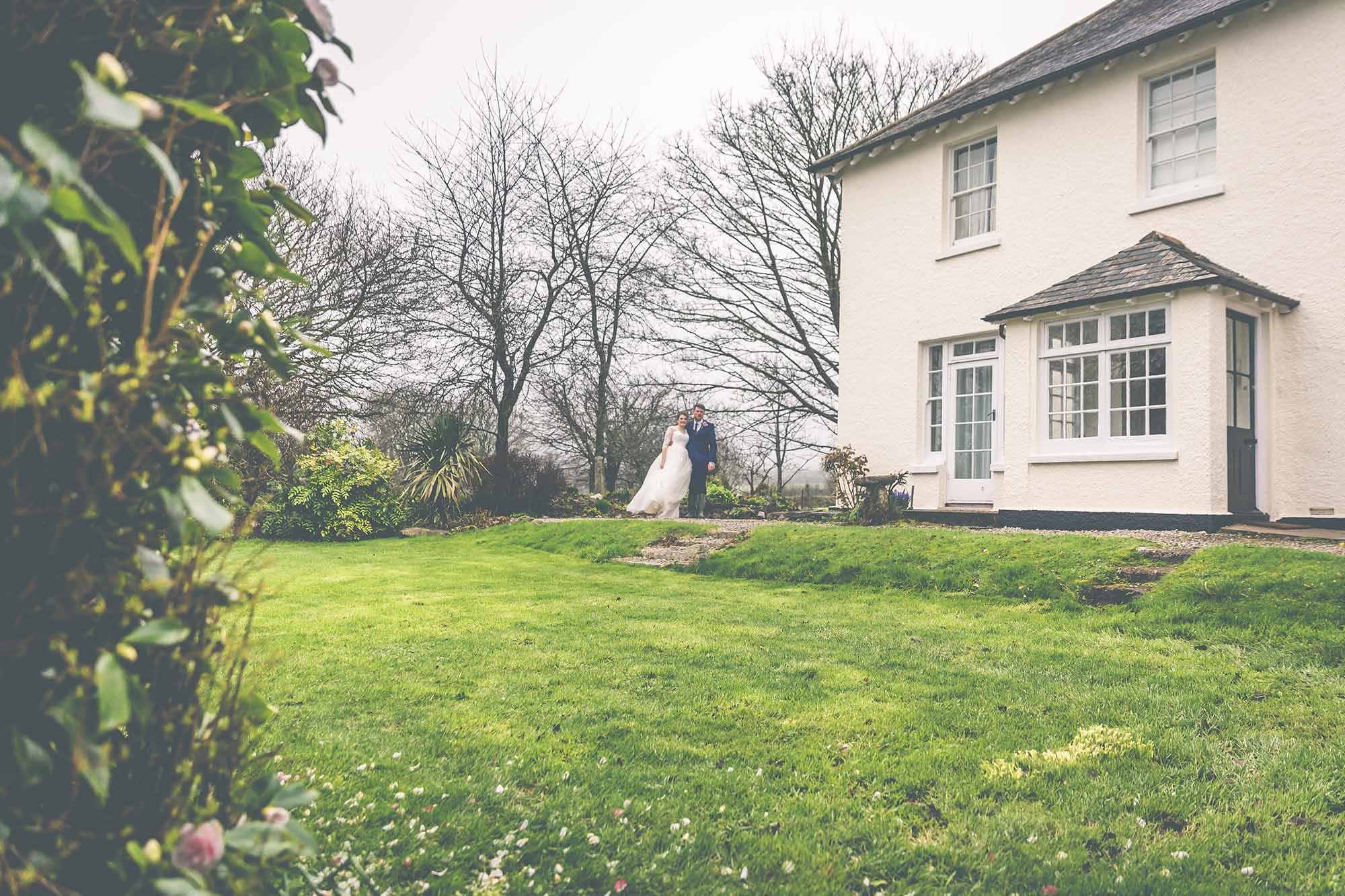 This is a picture of a bride and groom beside the farmhouse. The bride and groom are in the distance in the middle of the photo. The bottom half of the image is lawn. On the left there are some camellia bushes in the foreground. In the background there are some trees without any leaves. The farmhouse takes up the top, right hand quadrant of the image. Only part of the framhouse is in the shot. The farmhouse is rendered and painted white. It has a gray slate roof. The windows are georgian style sash windows and are painted white. There are two windows on the first floor and a French door on the ground floor. There is a porch with two sets of Georgian windows and a pitched slate roof. On the right hand side of the porch there is a black half-glazed door. There is a slight rise up towards the house and there is a small set of three steps on the left and a small step for three steps on the right of this ridge. The bride and groom are standing at the top of the steps on the left hand side of the house. The bride is wearing a white full gown with long sleeves. The groom is wearing a dark suit. There are some camellia petals on the lawn from the bushes indicating that the picture is taken in early spring.