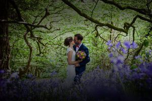 Newly weds kissing in a bluebell wood. The couple are in the centre of the image. They are surrounded by trees in leaf and there are lots of bluebells in the foreground. The image is taken in springtime. The couple are kissing the bride is on the left of the photo and the groom is on the right. The bride is wearing a lacy sleeveless dress you cannot see the couple's feet. In her right hand the bride is holding a bouquet of bright pink and bright yellow flowers. She has dark hair and she wears it up. The groom is wearing a mid blue suit with a white shirt and red bowtie. The groom has dark hair and a beard. The bride also has dark hair.