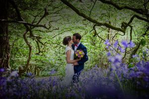 Newly weds kissing in a bluebell wood. The couple are in the centre of the image. They are surrounded by trees in leaf and there are lots of bluebells in the foreground. The image is taken in springtime. The couple are kissing the bride is on the left of the photo and the groom is on the right. The bride is wearing a lacy sleeveless dress you cannot see the couple's feet. In her right hand the bride is holding a bouquet of bright pink and bright yellow flowers. She has dark hair and she wears it up. The groom is wearing a mid blue suit with a white shirt and red bowtie. The groom has dark hair and a beard. The bride also has dark hair.