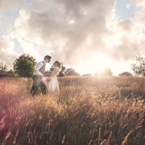 This is a summer wedding. The image is of the bride and groom in a field of long grass. The grass is parched fromthe summer sun. The couple are in the mid distance and it is a full length shot. They are slightly located off centre to the left of the photograph. Half of the images is of the grass and the top the top half is of the sky which is cloudy. There is a line of trees in the middle of the photo running accross the horizon. The bride and groom are facing each other and holding hands. The groom is leaning towards the bride. The groom wears dark trousers and a brown waistcoat. He wears a white shirt and a dark tie he has dark hair. The bride is wearing a white, full length dress. She has dark blond hair which she wears up loosely and is looking towards the groom.