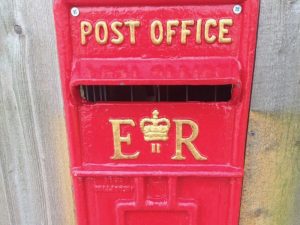 This image shows a wall-mounted traditional, metal, post-office postbox. It is red in colour with gold lettering. At the top it says "post office" and under the posting slot it says "E R". Between the E and the R there is a crown motif and a number 2. The postbox is mounted on a wooden wall.