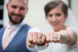This shot shows a bride and groom with their fists clenched showing off their wedding rings. The couple's fists are in focus and they are punching towards the camera. Their faces are blurred. The groom is on the left and the bride is on the right. The groom has dark here has a beard. he is wearing a mid-blue waistcoat, a white shirt and a pink tie. The bride is wearing a white dress with short sleeves and lace details and is wearing a veil. The image shows the couple from the chest up.